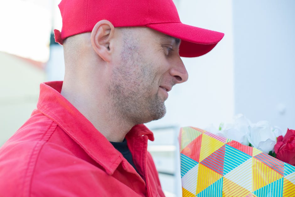 A smiling male flower delivery courier wearing a red cap and high-visibility vest extends a bouquet of fresh white roses and greenery towards the camera. The bouquet is wrapped in clear plastic and he