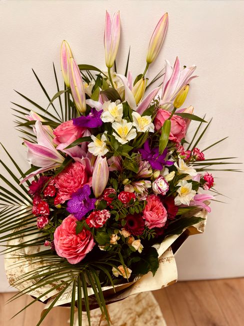 A large, vibrant floral arrangement featuring pink lilies with closed buds, pink carnations, white daffodils with yellow centers, and purple orchids, all nestled among lush green foliage including pal